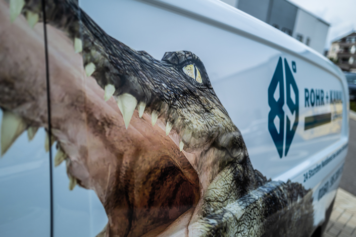 Nahaufnahme eines Fahrzeugs mit einem beeindruckenden Grafikdesign eines Krokodils. Das Auge des Krokodils und seine scharfen Zähne sind prominent dargestellt, während im Hintergrund das Logo und der Name "ROHR&KANAL" auf dem Fahrzeug zu sehen sind.