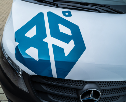 Nahaufnahme eines weißen Mercedes-Benz Fahrzeugs mit einem blauen Symbol auf der Motorhaube. Das Symbol zeigt zwei stilisierte Hammer-Silhouetten, die sich kreuzen, und eingebettete Diamantformen. Die Fahrzeugscheinwerfer und der Mercedes-Benz Stern sind ebenfalls sichtbar. Das Design des Fahrzeugs und das Symbol deuten auf eine professionelle und hochwertige Marke oder Dienstleistung hin.
